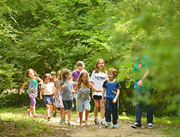 a group of children dressed in summer clothes walking on a dirt trail surrounded by grass and trees